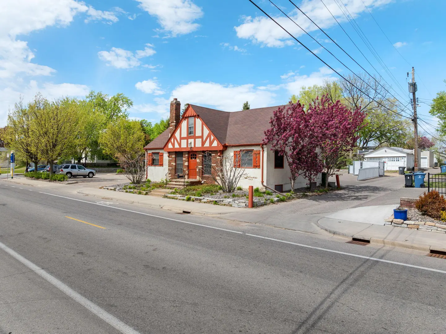 A charming Tudor-style house with red trim, shutters, and a brown roof sits on a corner lot with blooming trees under a blue sky.