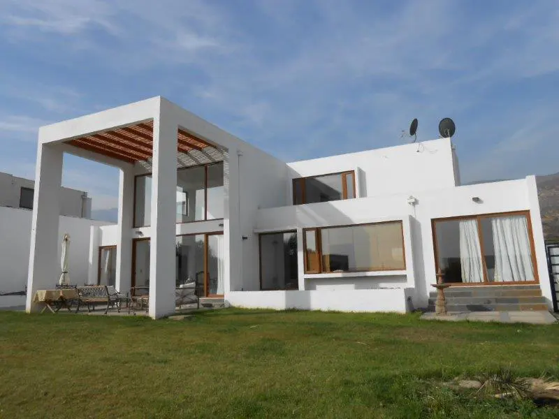 Modern white two-story house with large windows and a pergola, set on a green lawn under a blue sky.