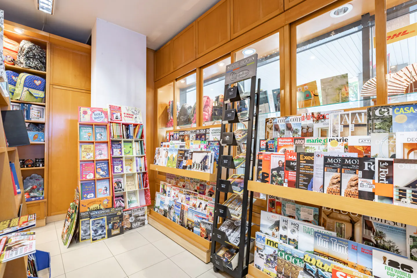 A bookstore interior with wooden shelves displaying books, magazines, and notebooks. A black metal display stand is in the center.