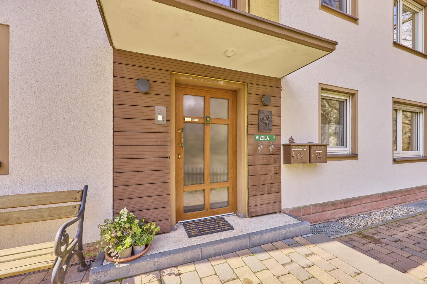 Exterior shot of a building with a wooden door, mailbox, and a bench with flowers.