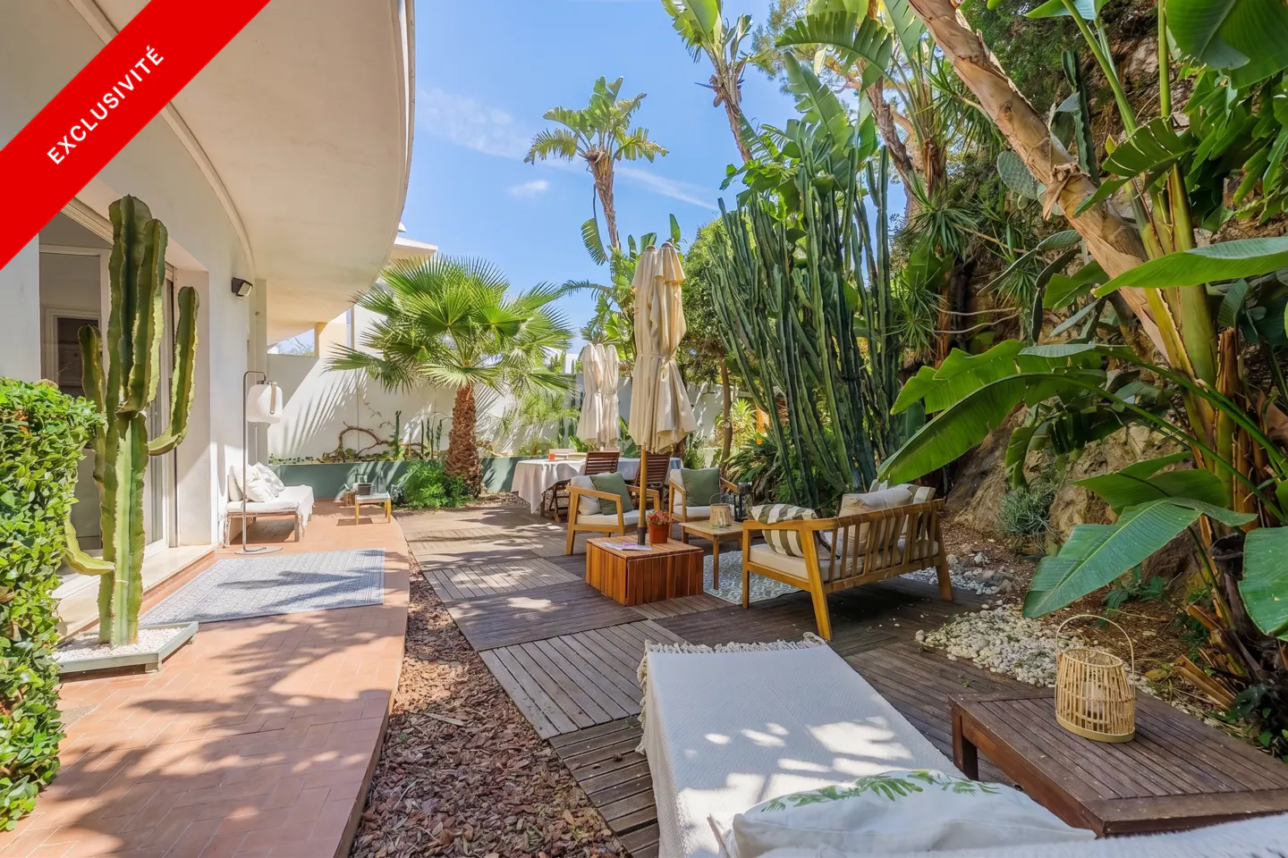 Outdoor patio with wooden furniture, palm trees, and lush greenery. A red "Exclusivite" banner is in the upper left corner.