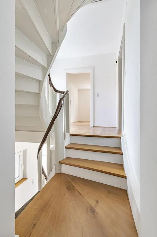 Interior view of a white spiral staircase with wood steps and a brown handrail leading to a doorway.