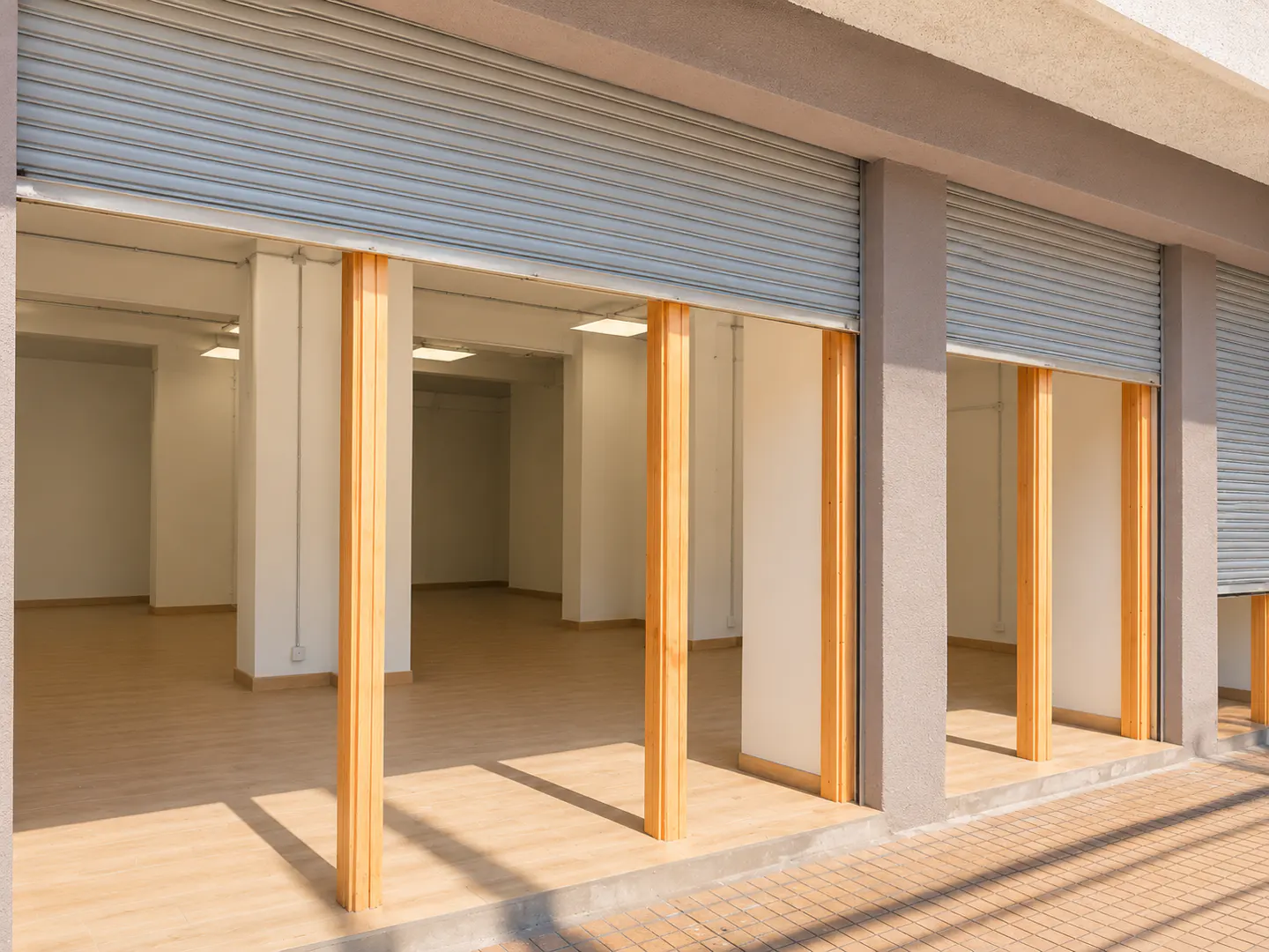 Empty commercial storefront with light wood flooring, white walls, and vertical wooden pillars under metal security shutters.