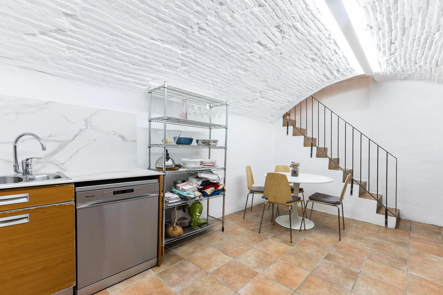 A kitchen area with a sink, dishwasher, metal shelving, table with chairs, and stairs. The floor is tiled.