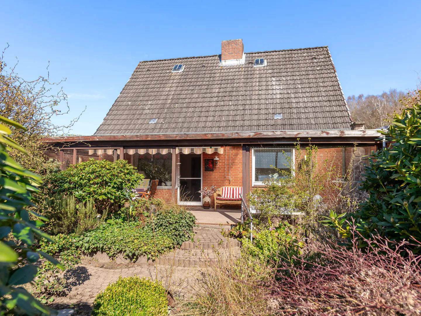 Backyard view of a brick house with a gray tiled roof, chimney, and skylights, surrounded by lush green plants.