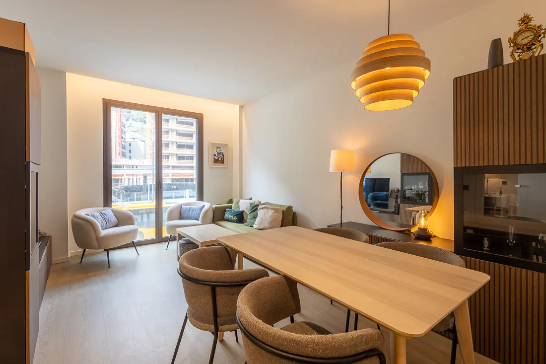 Bright living room with a light wood table and chairs, a green sofa, and a large window with a city view.