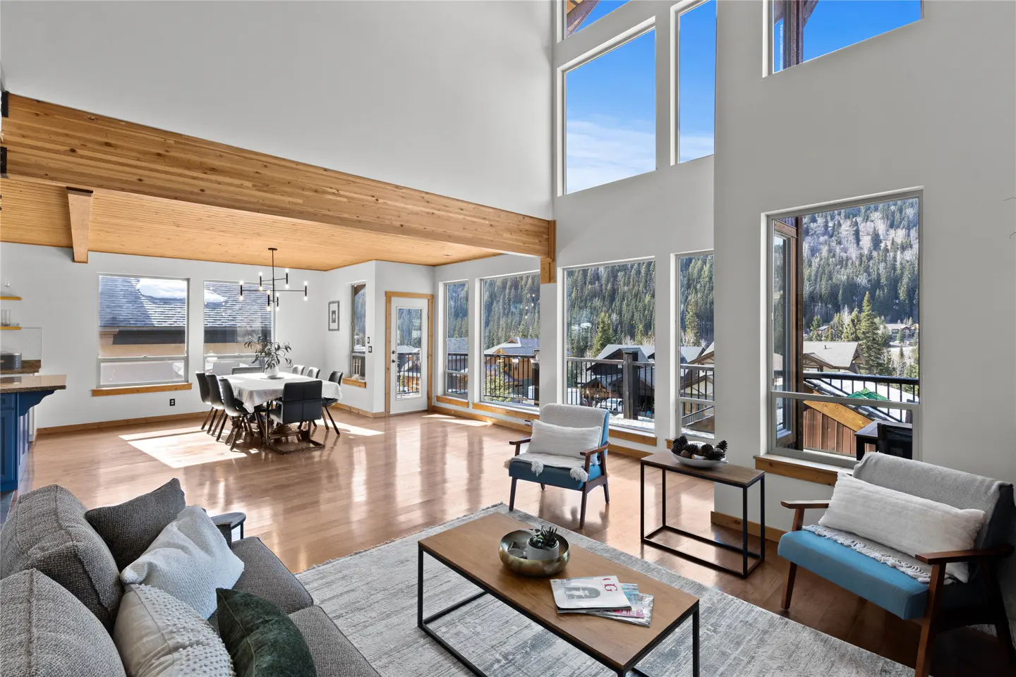 Bright, open living room with wood floors, large windows, and mountain views. Gray sofa, blue chairs, and dining table visible.