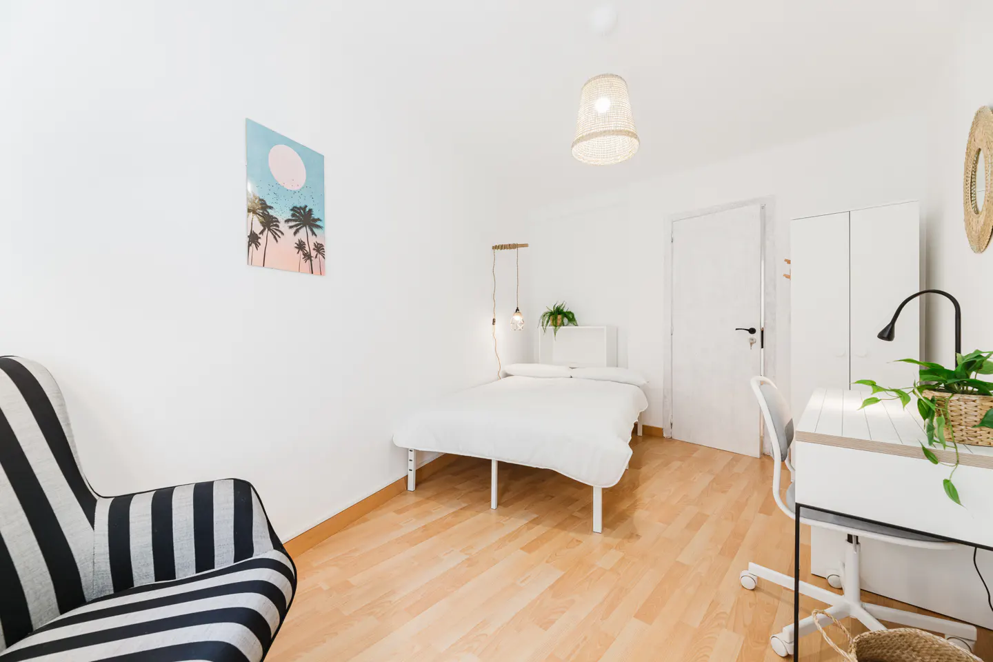 Bright bedroom with white walls, wood floors, and a black and white striped chair. A white bed, desk, and palm tree art add to the room's decor.