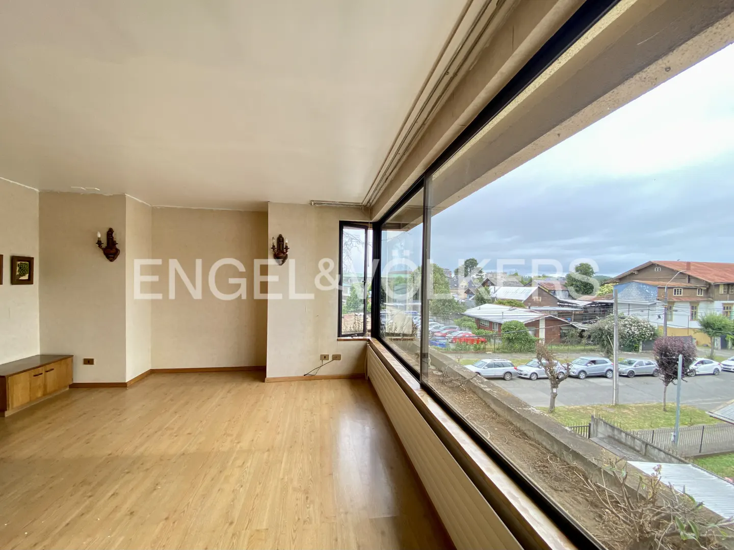 A bright, empty room with wood floors and a large window overlooking a street with houses and cars. The Engel & Volkers logo is visible on the wall.