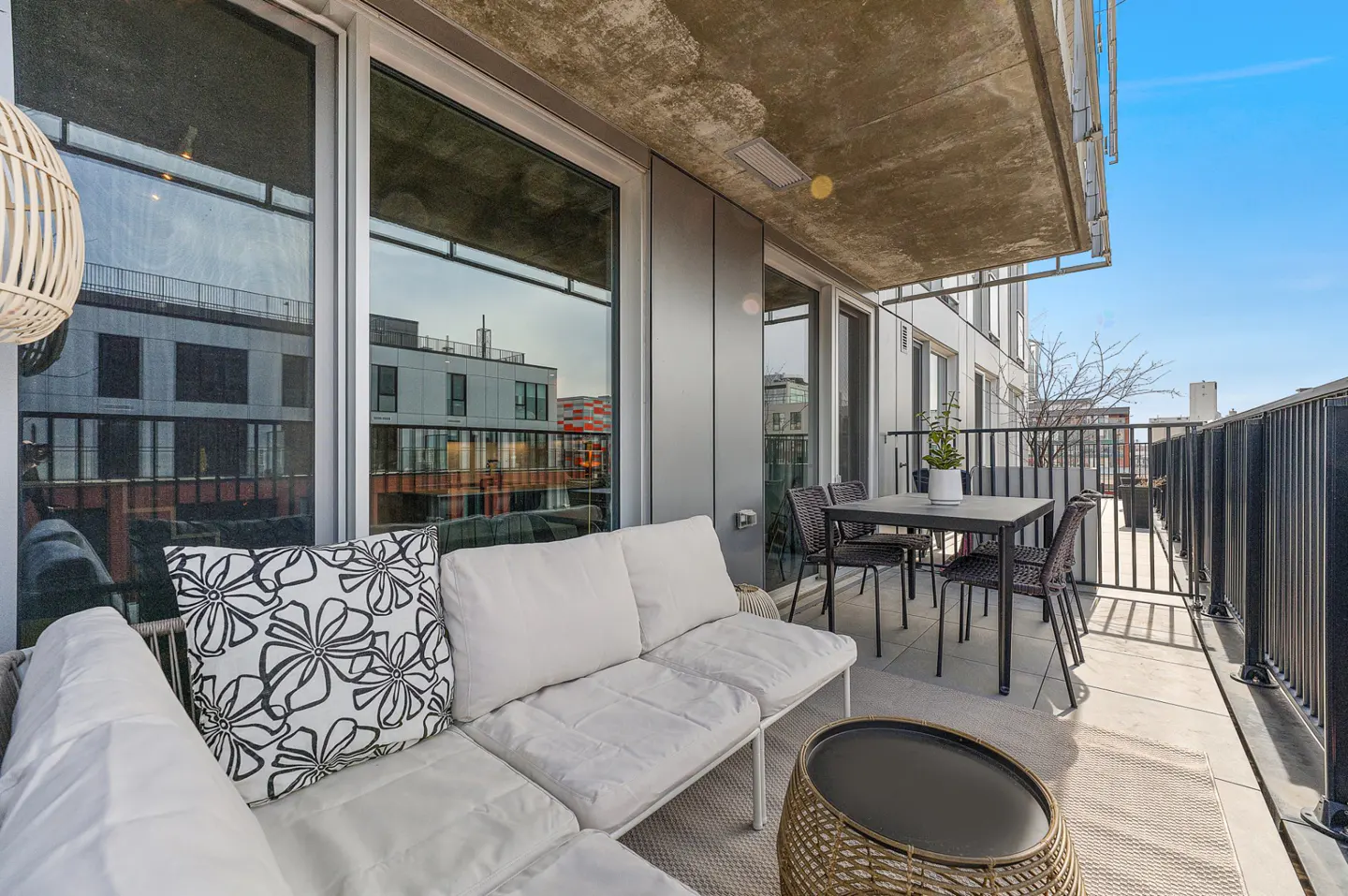 Balcony with white cushioned sofa, black table and chairs, and a wicker side table. Black metal railing and city views.