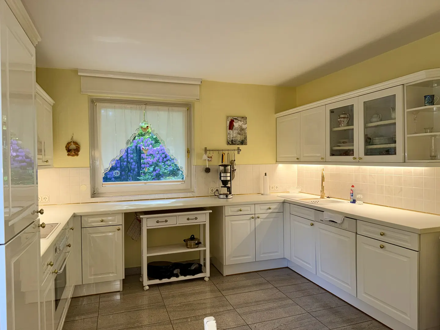 Bright kitchen with white cabinets, yellow walls, tiled floors, and a window view of purple flowers.