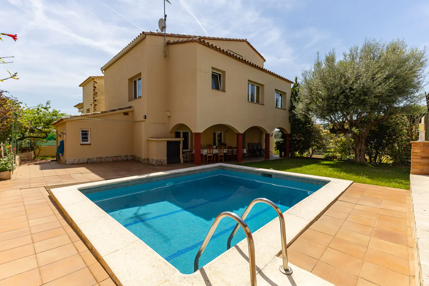 A two-story beige house with a red tile roof overlooks a blue tiled pool with a metal ladder. A covered patio and green lawn are visible.