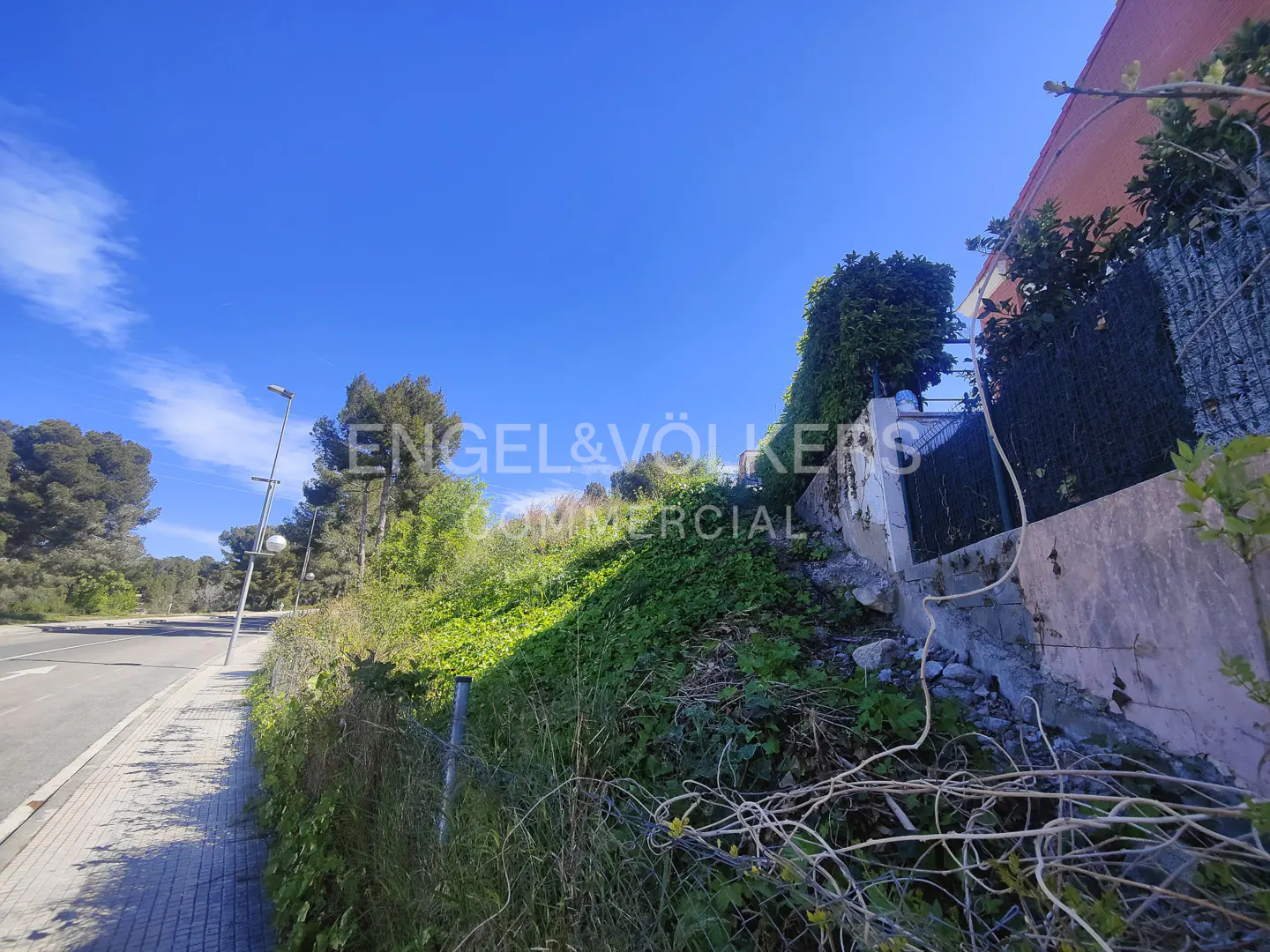 Exterior view of a grassy hillside lot next to a road, under a clear blue sky. The Engel & Völkers logo is superimposed on the image.