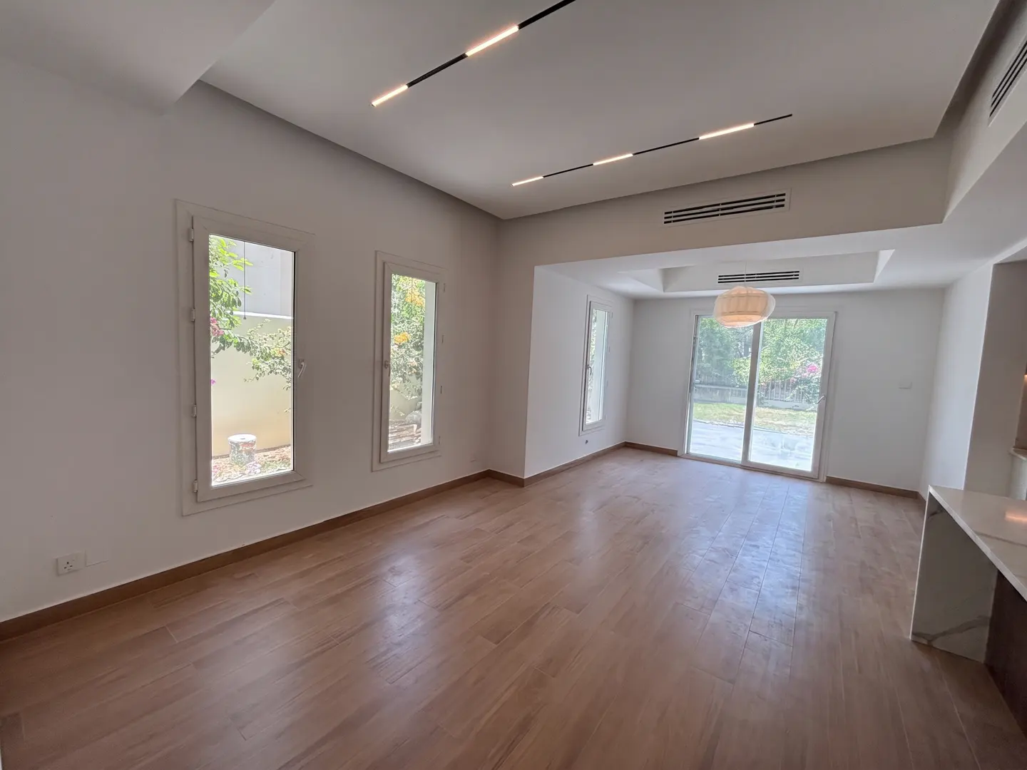 Bright, empty room with wood floors, white walls, and modern lighting. Windows and sliding glass doors offer views of greenery.