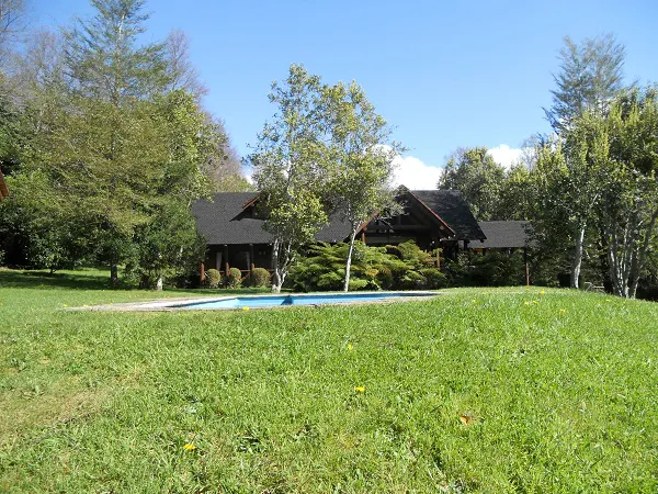 A log cabin with a black roof and a blue pool is surrounded by green grass and trees.