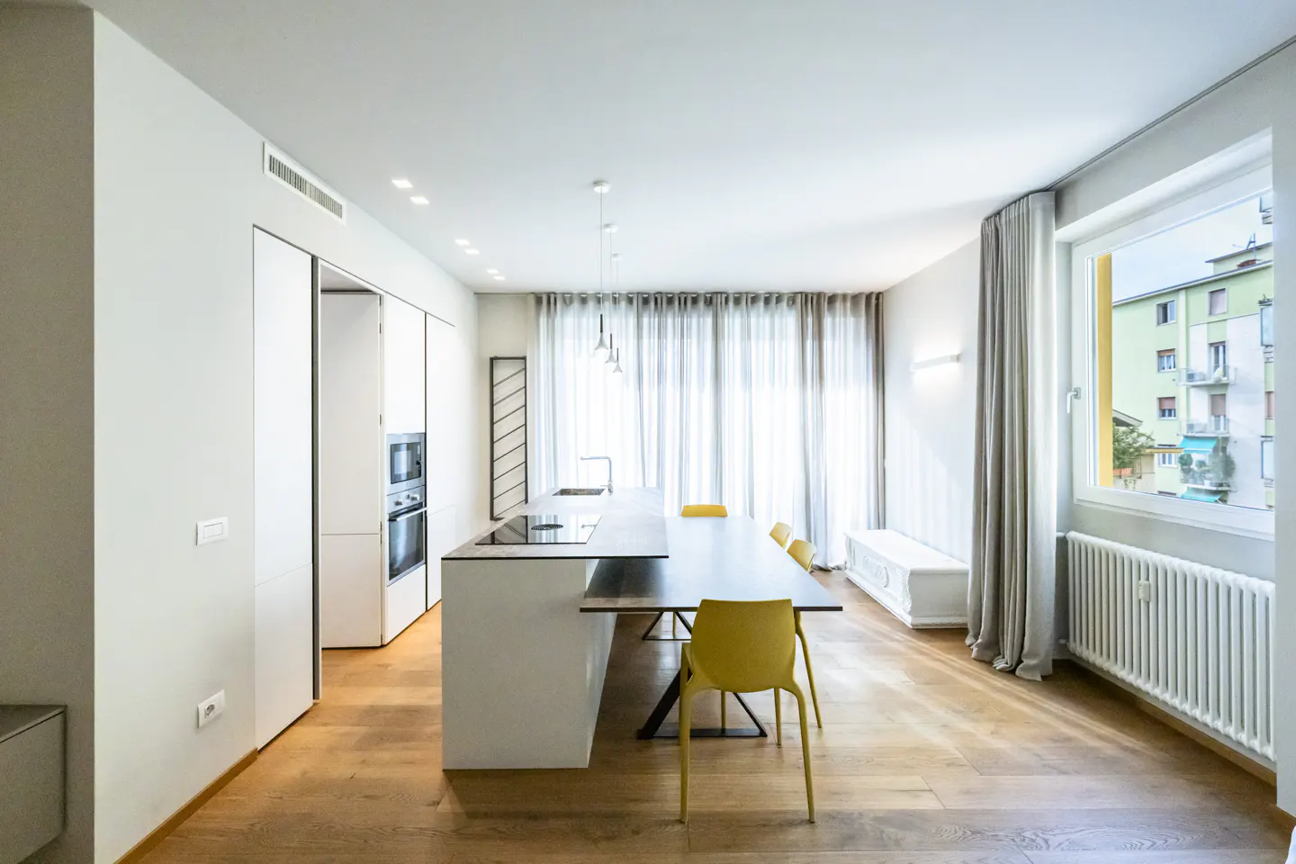 Bright, modern kitchen with white cabinets, a black island, and yellow chairs. A large window with sheer curtains lets in natural light.