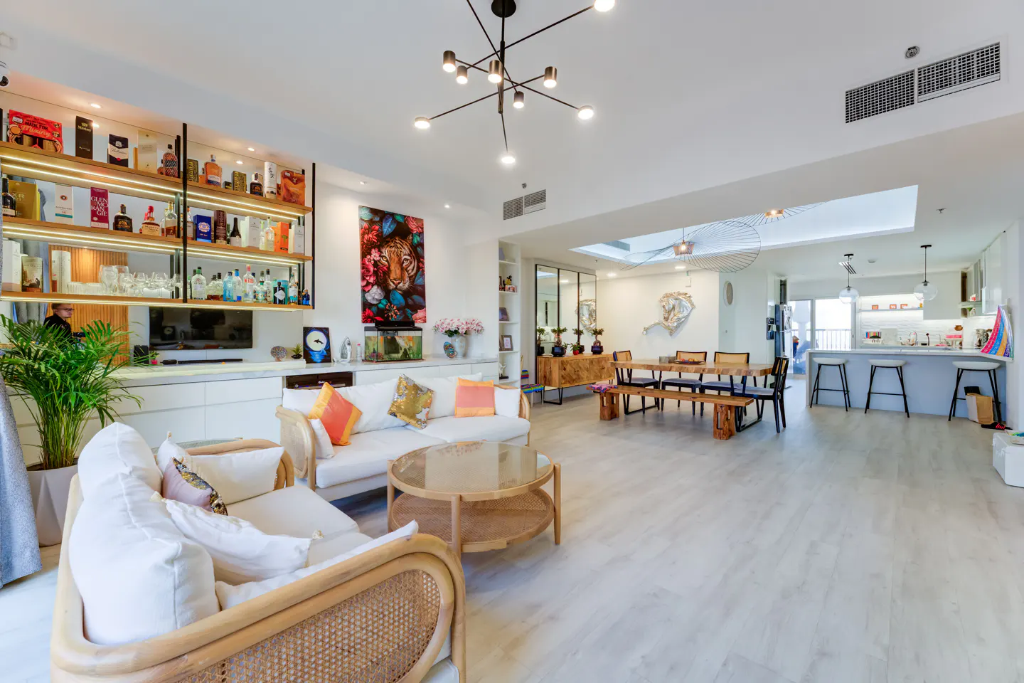 Bright, open-concept living space with white walls, light wood floors, and modern furnishings. A bar area, dining table, and kitchen are visible.