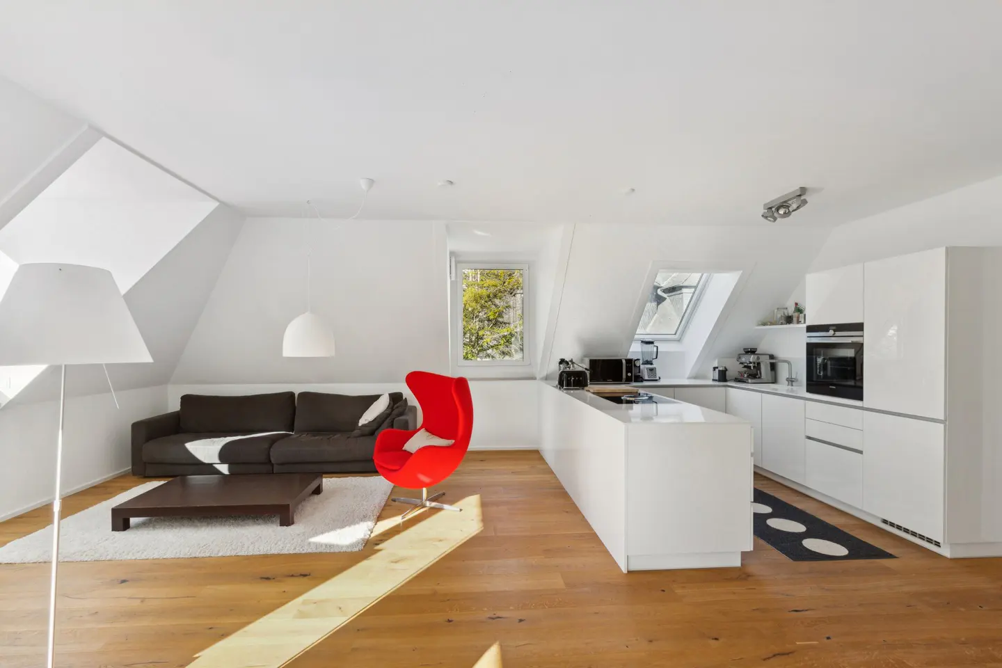 Bright, modern attic apartment with wood floors, white walls, and a brown sofa. A red egg chair sits near a white kitchen with skylight.