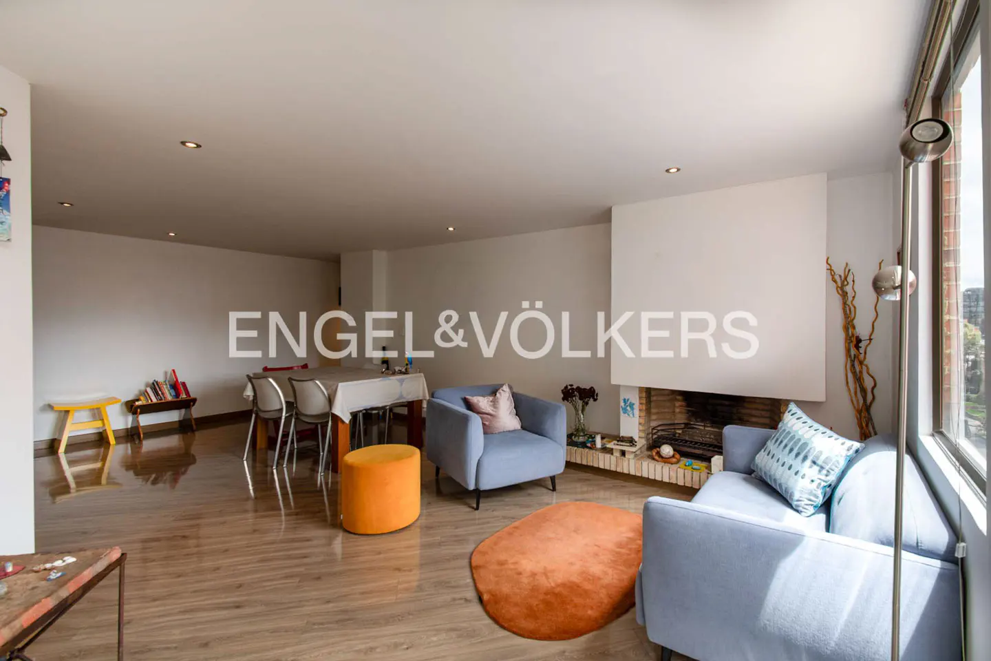 Bright living room with wood floors, blue sofas, orange rug, and fireplace. Dining table and chairs in the background. Engel & Völkers logo visible.