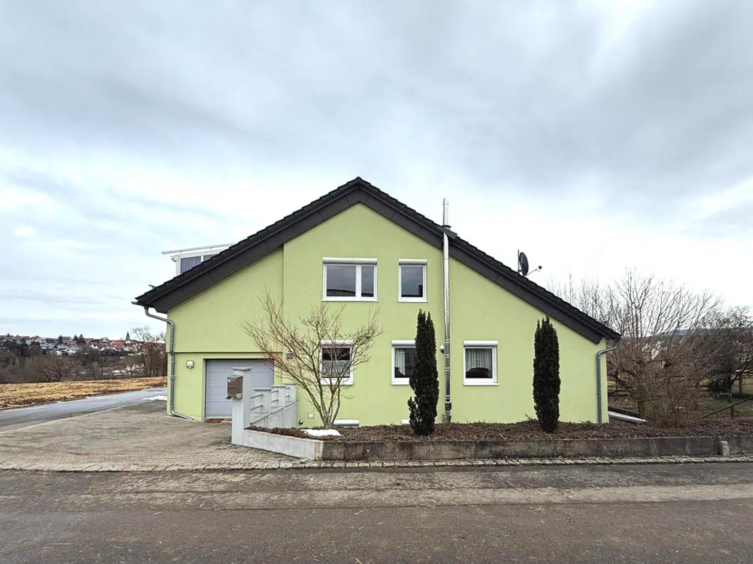 Two-story light green house with a dark roof, white windows, and a silver chimney on a cloudy day.