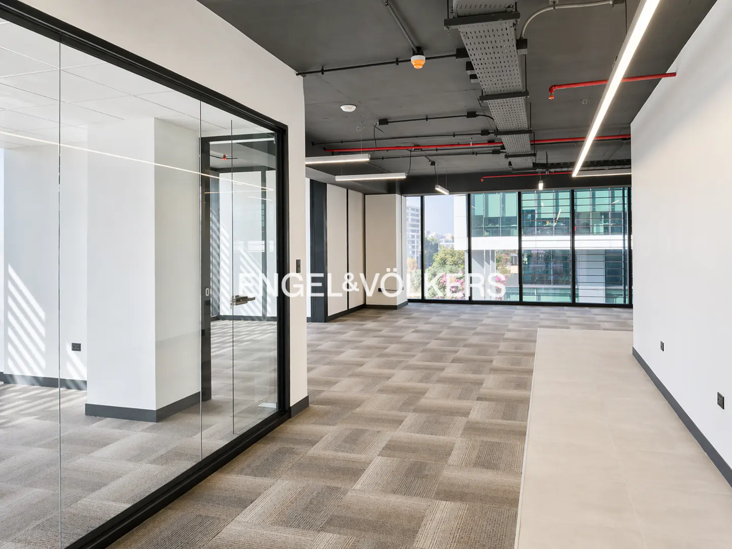 Bright, empty office space with gray carpet, white walls, and floor-to-ceiling windows. The ceiling is exposed with visible pipes and wiring. Engel & Völkers logo on glass wall.