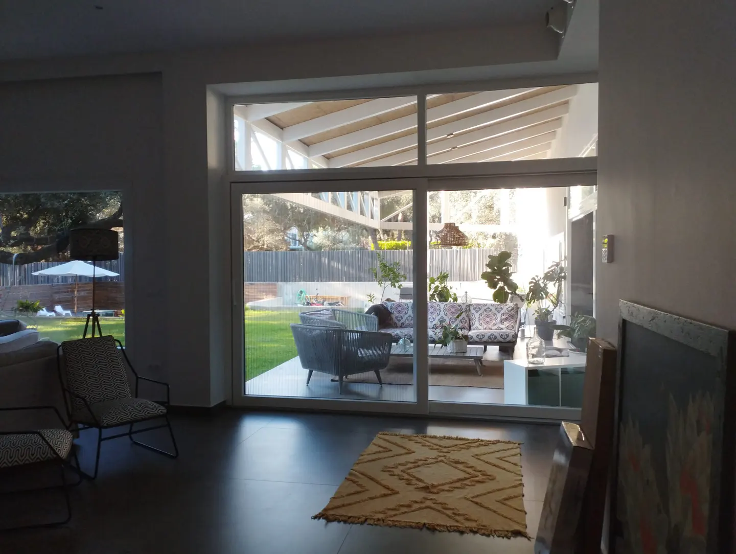 A bright living room with a large sliding glass door to a patio with outdoor furniture. A patterned rug sits on the dark floor.