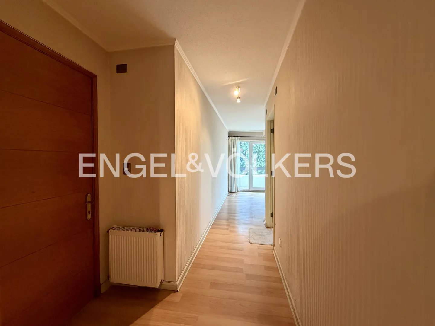 Hallway view with wood floors, beige walls, and a brown door on the left. A radiator is visible near the door. Light streams in from the end of the hall.