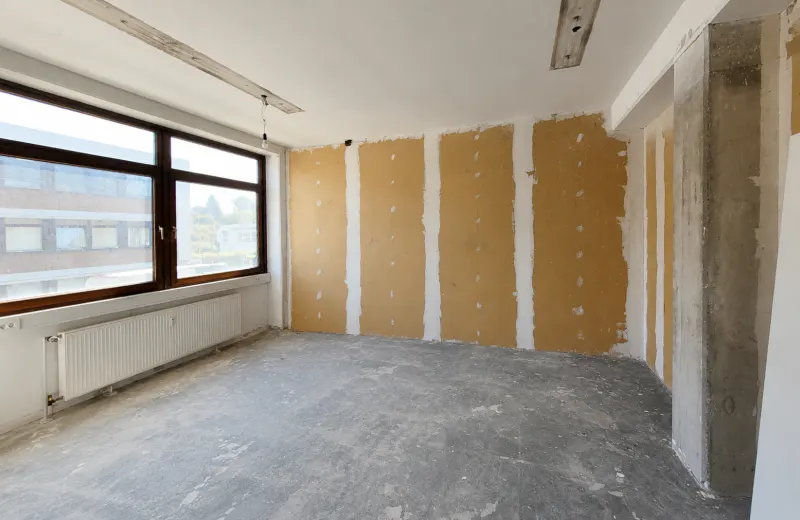 An unfinished room with bare concrete floors, walls with exposed drywall seams, a large window, and a white radiator.