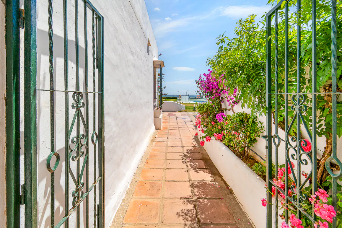 View through an open green iron gate to a terracotta tiled path, white wall, and colorful flowers. Blue sky in the background.