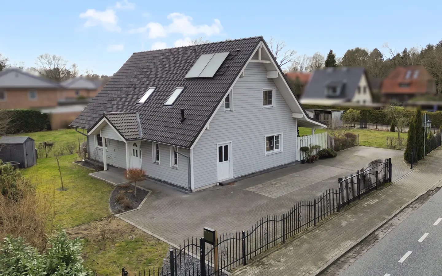 A two-story light gray house with a dark gray roof, solar panels, and a brick driveway. A black metal fence borders the property.