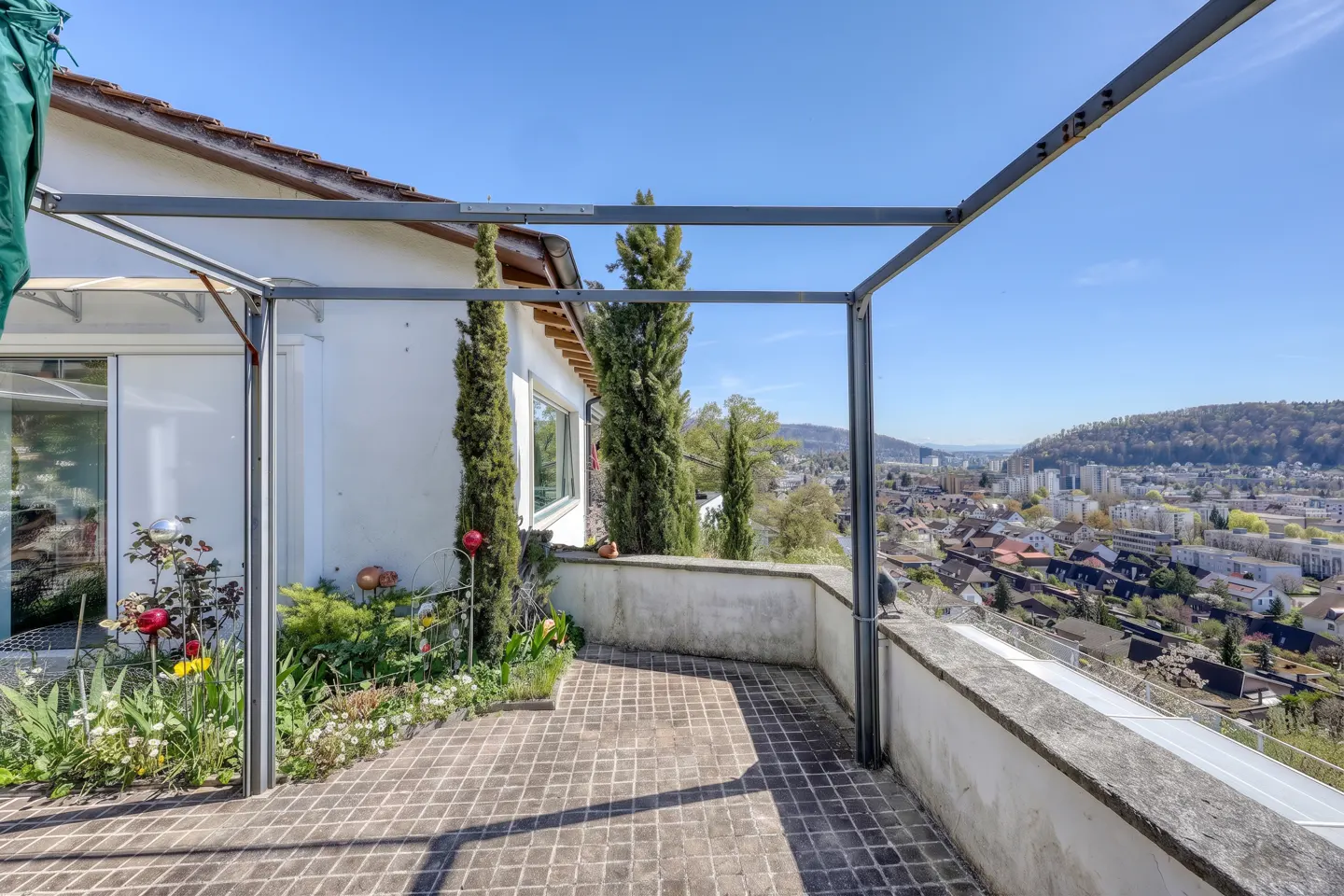 A patio with a metal pergola overlooks a city. The patio has a brick floor, a low wall, and a garden with flowers.