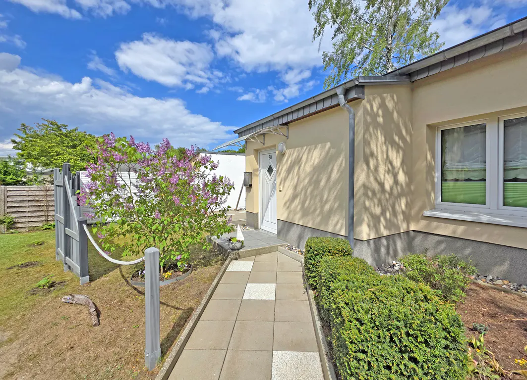 A beige house with a white front door, a tiled walkway, a blooming lilac bush, and green hedges under a blue sky.