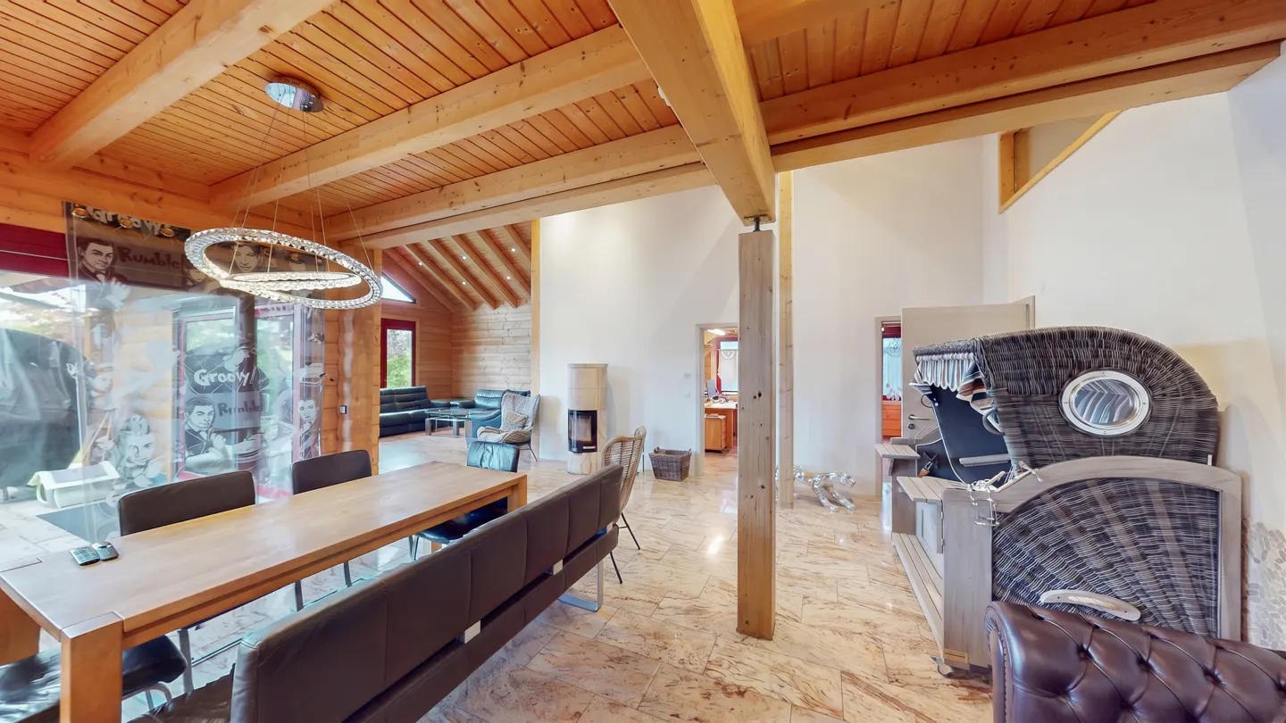 Open-concept living space with wood-beamed ceilings, a dining table with a bench, and a unique wicker chair.