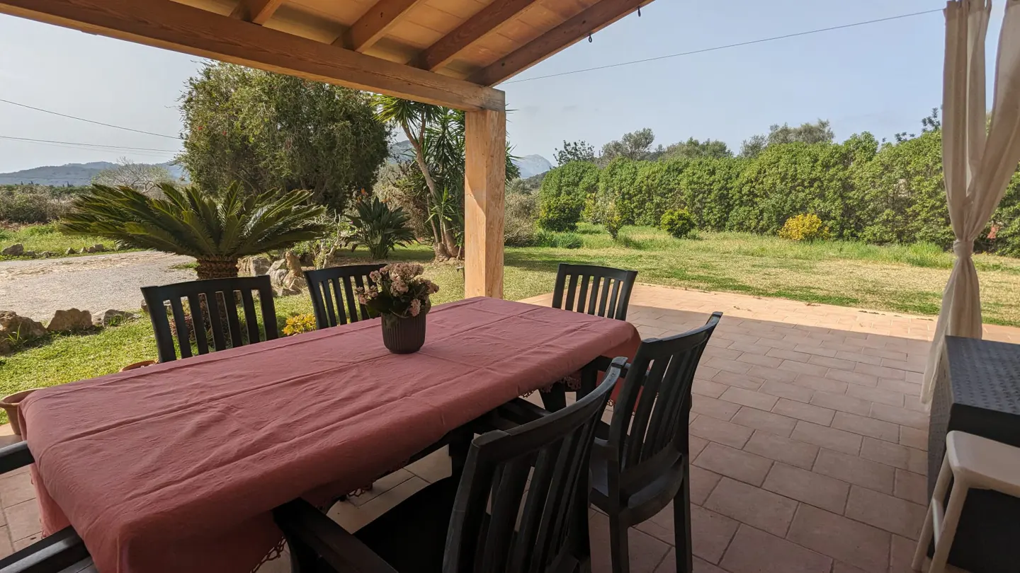 Outdoor patio with a table covered in a red cloth, surrounded by black chairs. A vase of flowers sits on the table, with a green lawn and trees in the background.