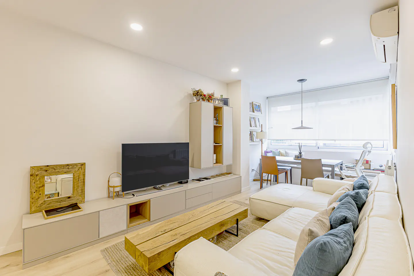 Modern living room with a white sectional sofa, blue pillows, a wooden coffee table, and a wall-mounted TV unit.