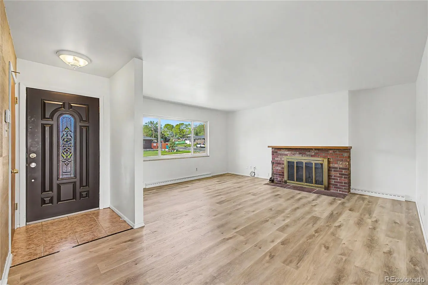 A bright living room with wood floors, white walls, and a brick fireplace. A front door with stained glass is visible.