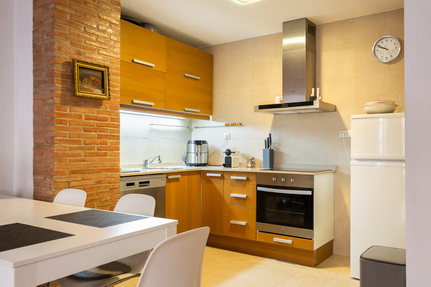 A bright kitchen with wooden cabinets, stainless steel appliances, and a white table with chairs. A brick column with a framed picture adds character.
