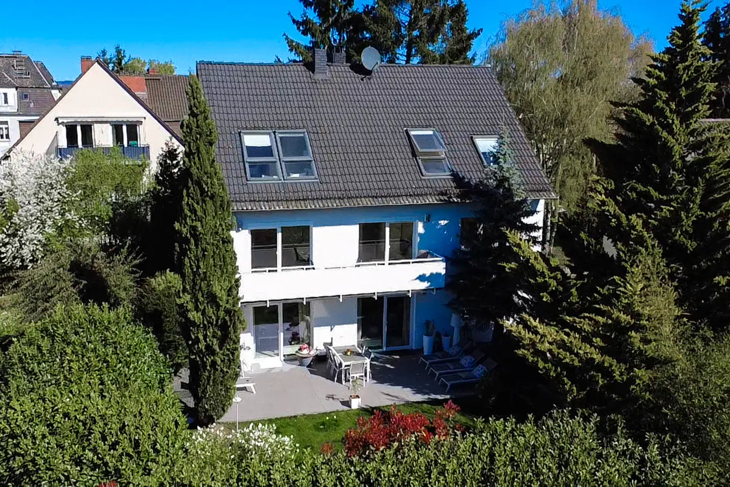 Two-story white house with a gray roof, skylights, and a patio with outdoor furniture. Green trees and bushes surround the house.