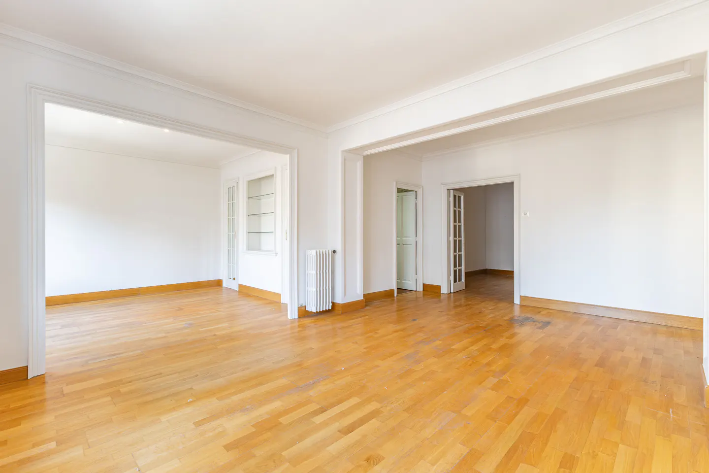 Bright, empty room with hardwood floors and white walls. Doorways lead to other rooms. A radiator and built-in shelving are visible.