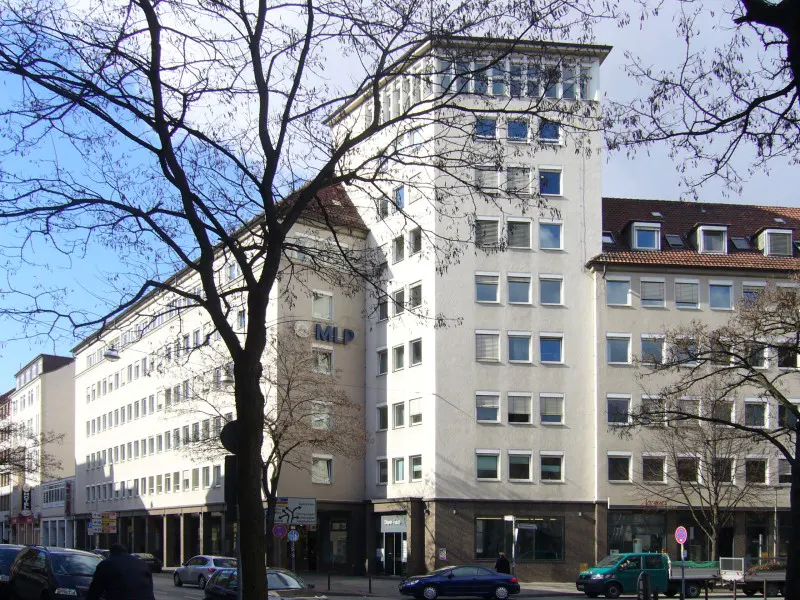Exterior view of a multi-story white building with many windows, cars parked on the street, and bare trees in the foreground.