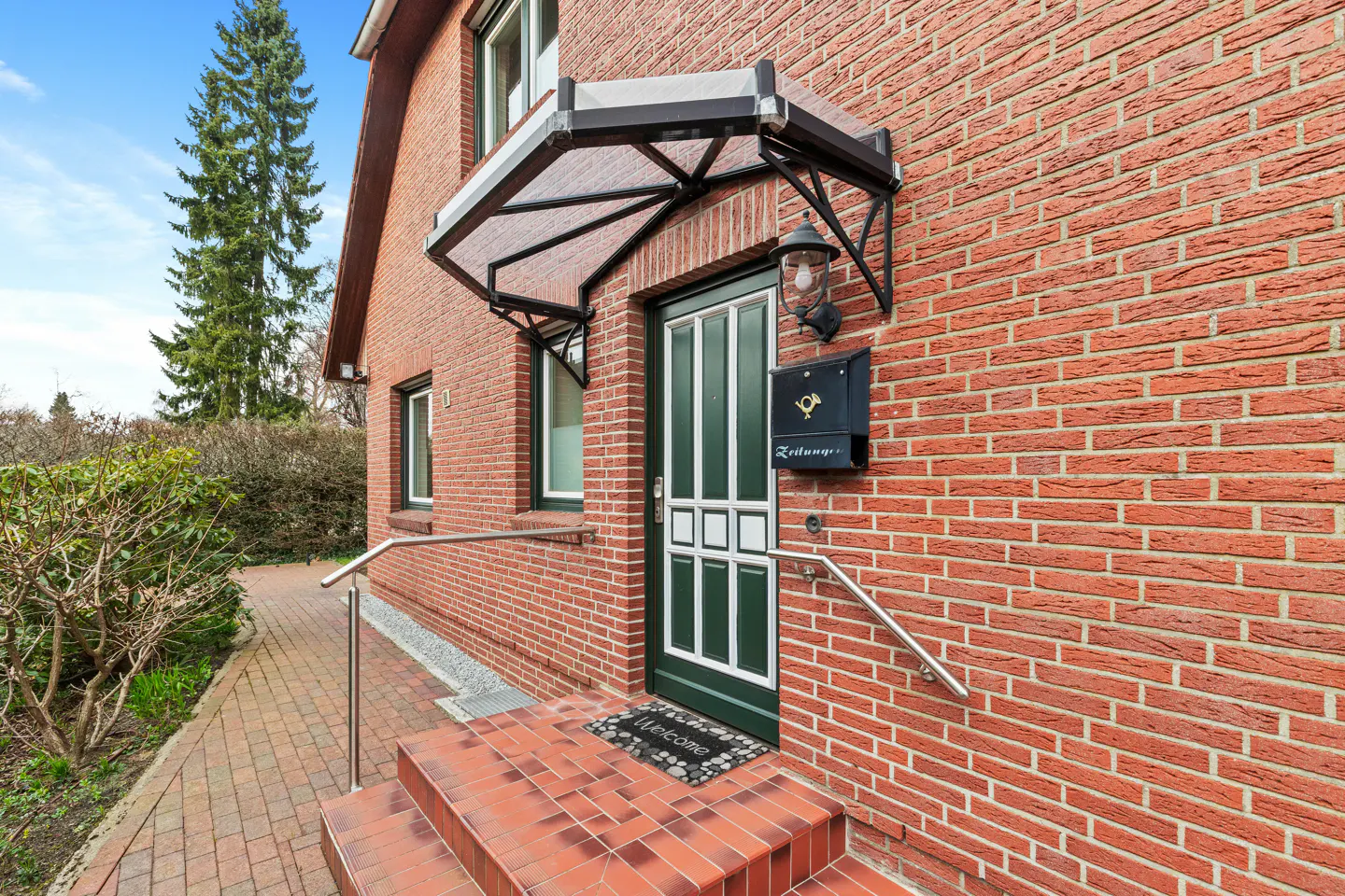 Brick house exterior with a green door, black mailbox, and clear awning. A "welcome" mat sits on the red tiled steps.