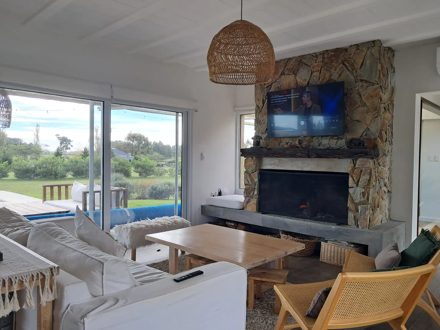Living room with white sofa, wood table, stone fireplace with TV, and large windows overlooking a green lawn.
