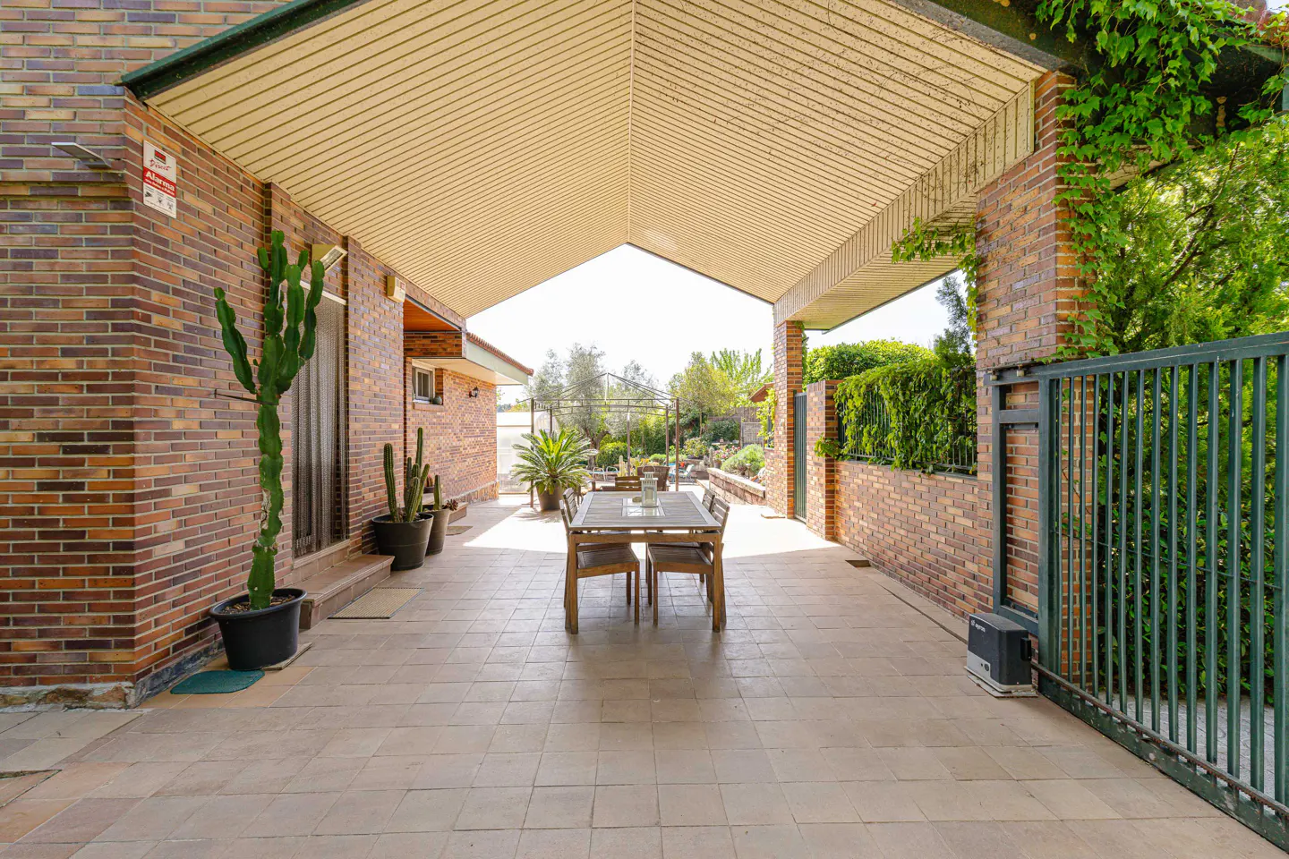 Covered patio with brick walls, tile floor, and a wooden table with chairs. Cacti in pots line the wall. A green metal gate leads to the garden.