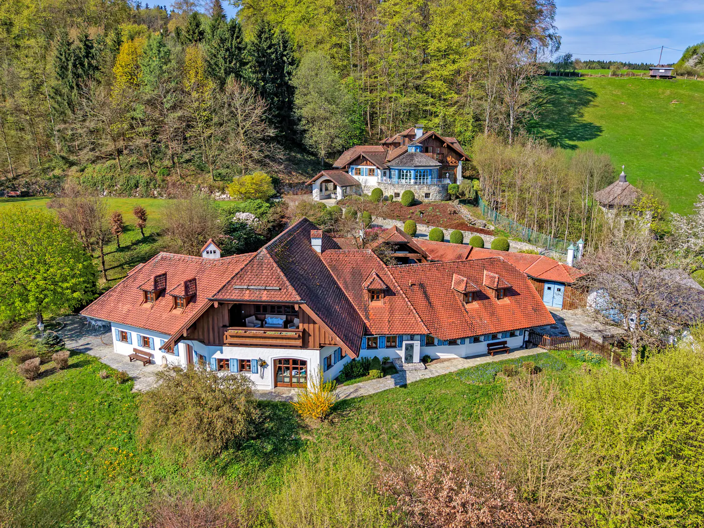 Aerial view of a large, white house with a red tile roof, blue shutters, and a balcony, surrounded by green trees and grass.