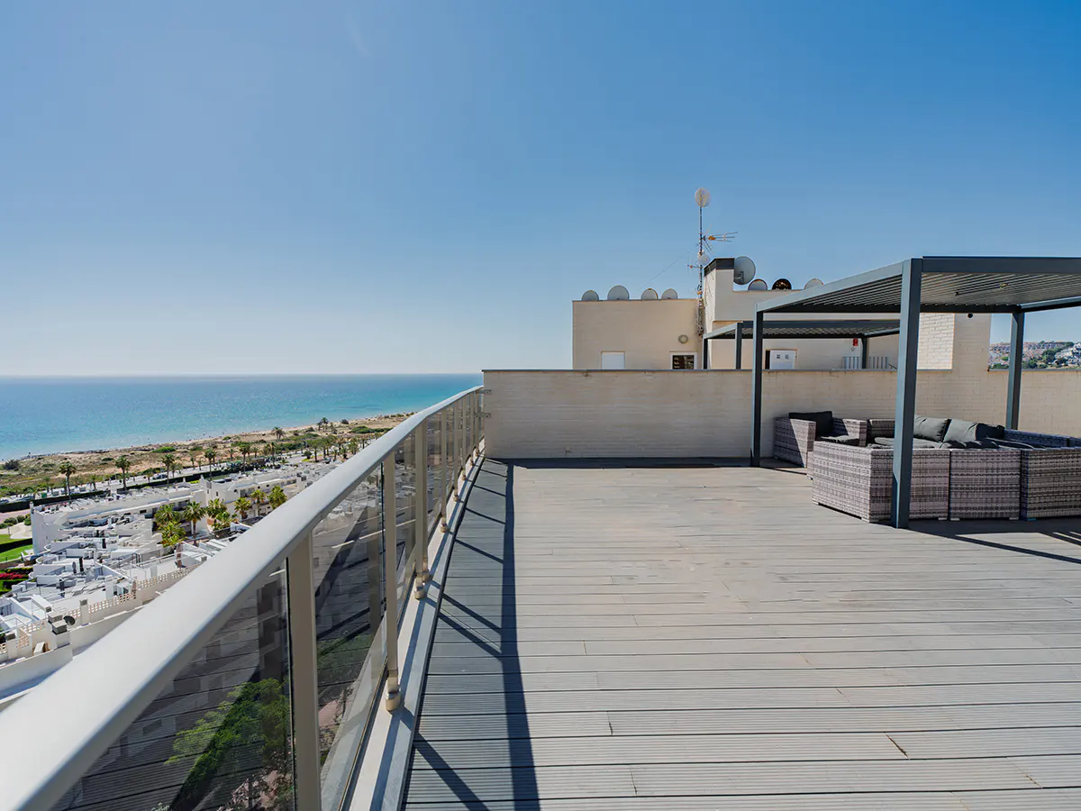 Rooftop patio with ocean view. Gray wicker furniture sits under a pergola. A glass railing lines the edge of the deck.
