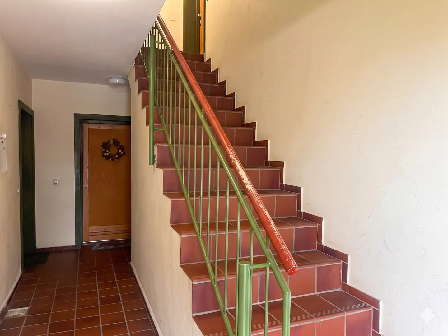 Hallway with terracotta tile floor and stairs. Green metal railing with red handrail. Door with wreath visible.