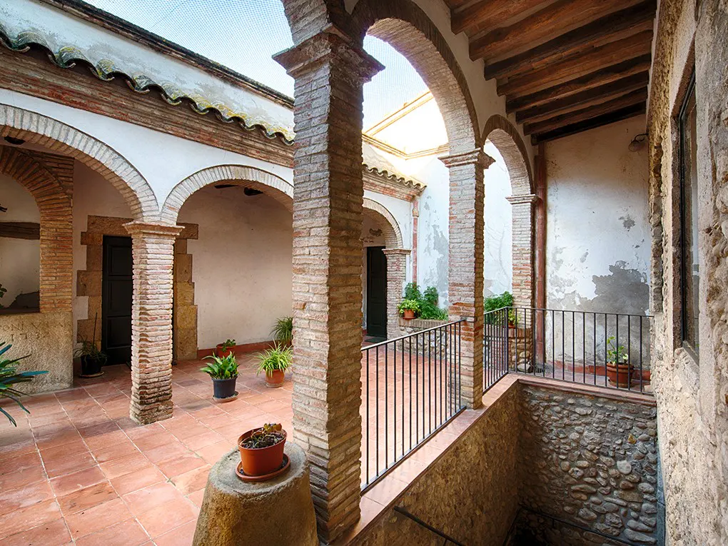 Courtyard with brick arches, columns, and terracotta tile floor. Potted plants are scattered throughout. A stone well is visible in the foreground.