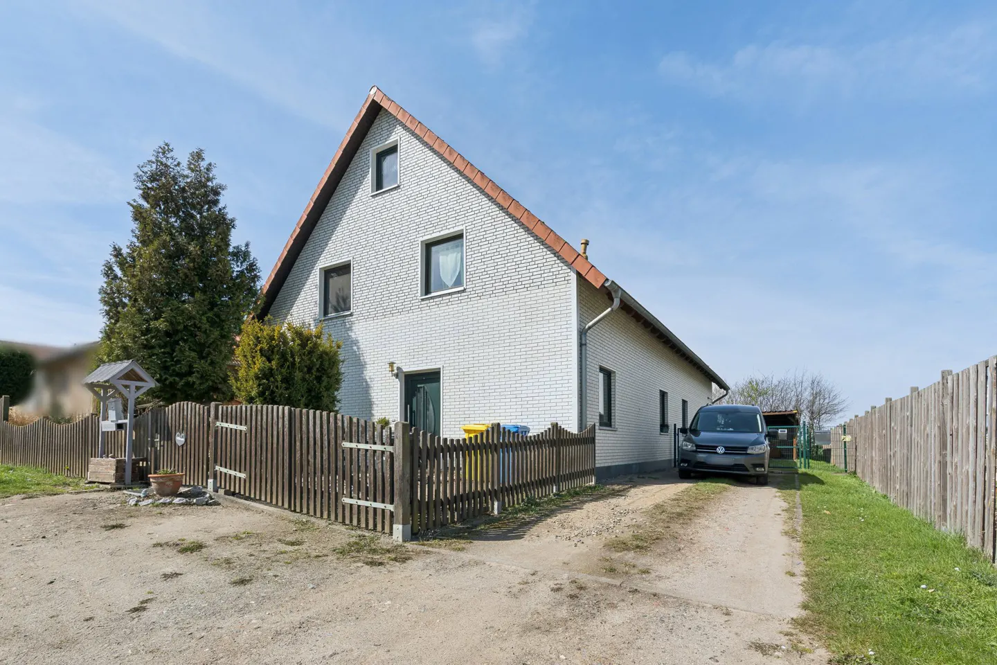 Two-story white brick house with a brown roof, a wooden fence, and a gray car parked in the driveway.