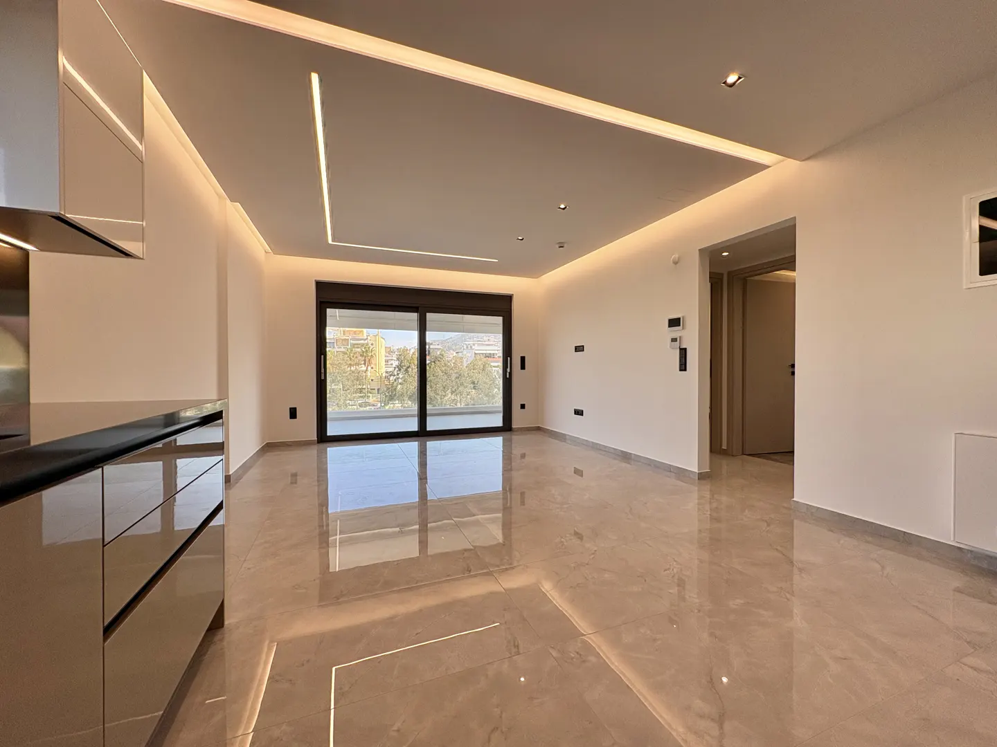 A modern apartment interior with beige cabinets, a black countertop, and a sliding glass door to a balcony.