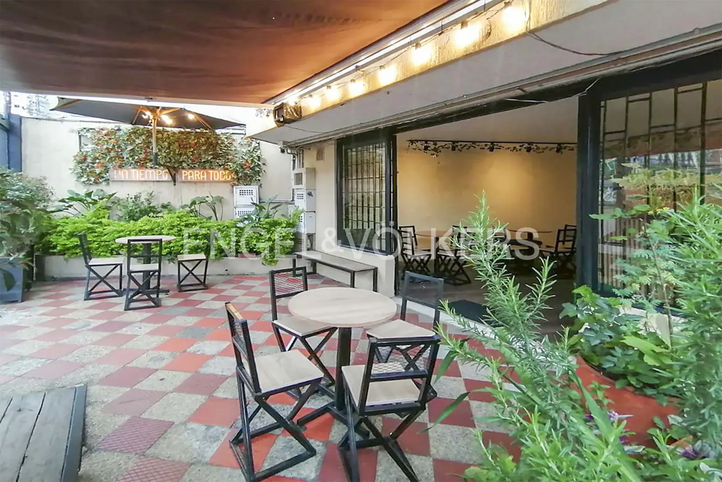Outdoor patio with tables, chairs, and greenery. The floor is tiled in a red and white checkered pattern. A retractable awning covers the space.